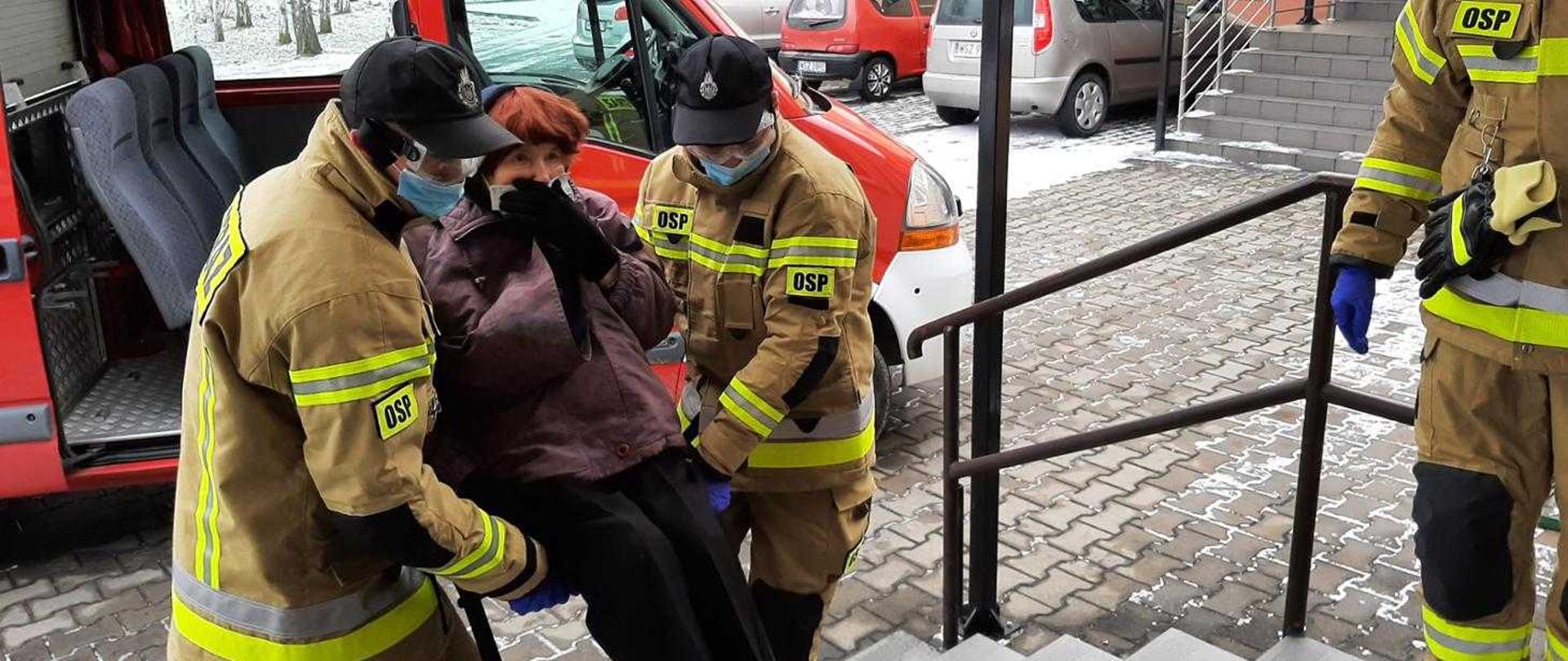 Strażacy wnoszący na rękach starszą kobietę na szczepienie przeciw COVID-19.