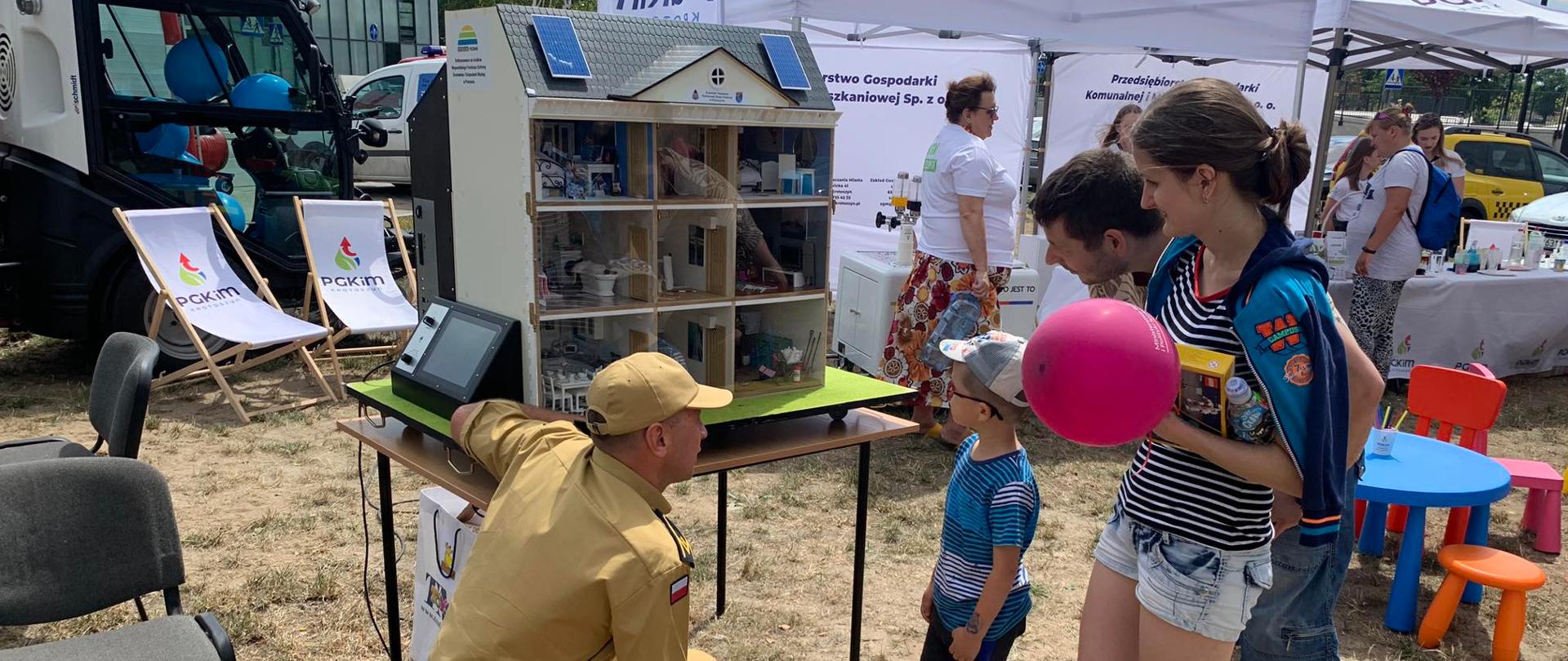 Podczas pikniku rodzinnego krotoszyńscy strażacy prezentują zagadnienia prewencji społecznej z użyciem mobilnego symulatora zagrożeń. Przy domku edukacyjnym grupa dzieci z opiekunami podziwia prezentacje zagrożeń omawianą przez strażaka.