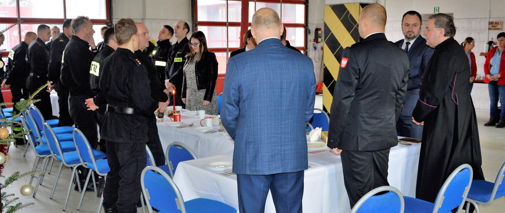Na zdjęciu widać starostę powiatu bieszczadzkiego wraz z zastępcą, komendanta powiatowego PSP w Ustrzykach Dolnych, księdza oraz strażaków z KP PSP w Ustrzykach Dolnych przy stole wigilijnym na garażu komendy. W tle bramy garażowe.