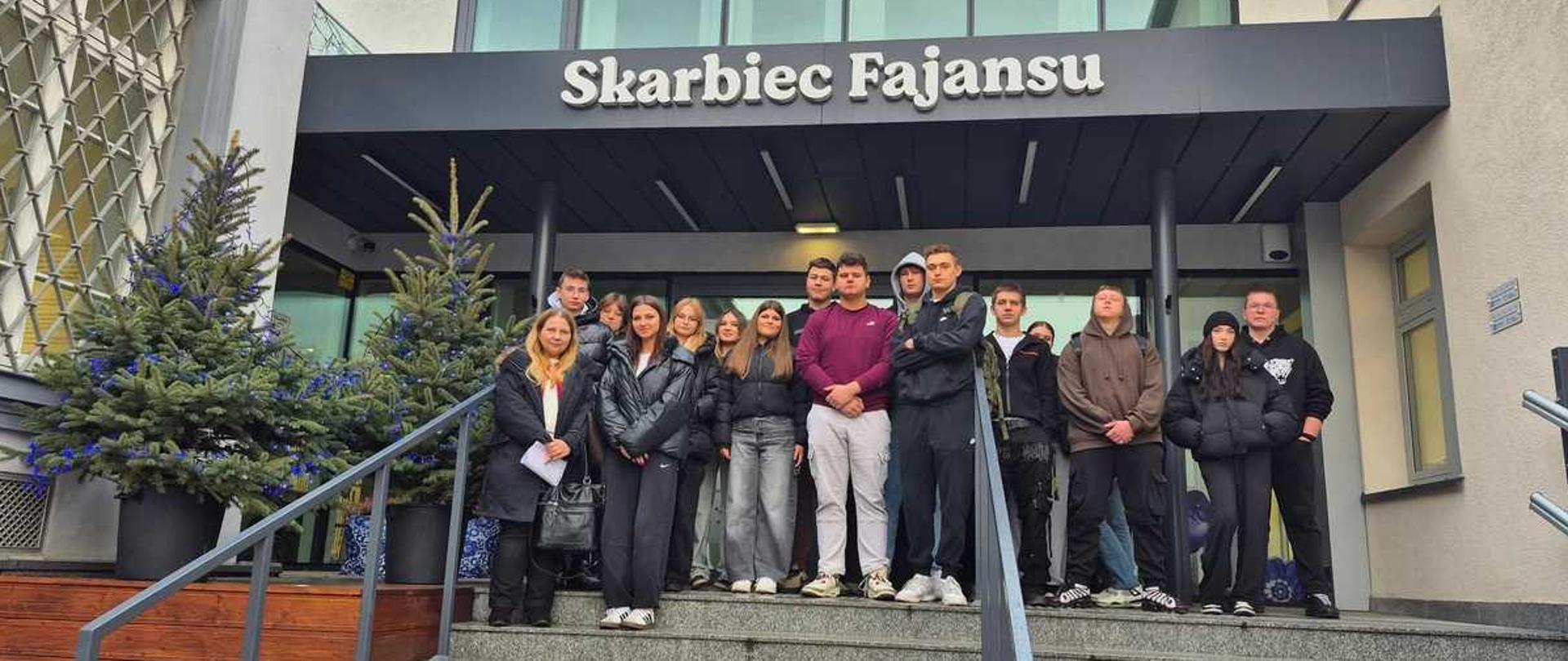 Zdjęcie przedstawia grupę kilkunastu młodych ludzi stojących na szerokich, zewnętrznych schodach prowadzących do wejścia budynku. Grupa składa się z młodzieży w wieku szkolnym, ubranej w różnorodne, codzienne ubrania, takie jak kurtki, bluzy i dżinsy. Tło stanowią nowoczesne budynki.
Centralnym punktem, widocznym bezpośrednio za grupą, jest główne wejście, nad którym znajduje się duży, czarny szyld z białym napisem: ""Skarbiec Fajansu"". Obiekt ten to Interaktywne Centrum Fajansu mieszczące się we Włocławku, będące filią Centrum Kultury ""Browar B."". Centrum to, otwarte w 2023 roku, ma na celu prezentację 150-letniej historii włocławskiej ceramiki poprzez multimedialne wystawy i warsztaty.
Schody wykonane są z szarego betonu, a z lewej strony znajduje się drewniana rampa lub podest. Po obu stronach wejścia, na tarasie, stoją duże donice z udekorowanymi choinkami, co sugeruje okres świąteczny. Po lewej stronie widoczna jest ozdobna, przeszklona fasada starszego budynku, a po prawej bardziej nowoczesna, jasnoszara elewacja.