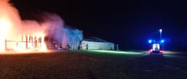 Na zdjęciu widać stodołę w całości objętą pożarem, samochód gaśniczy oraz strażaków podczas gaszenia pożaru.