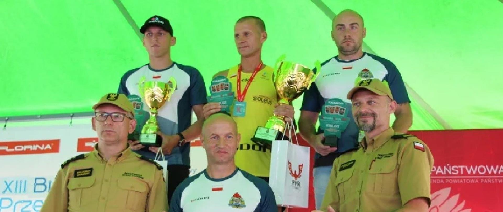Trzech mężczyzn w strojach sportowych stoi na podium, przed nimi stoi trzech mężczyzn, dwóch stojących po bokach ubranych jest w mundury straży pożarnej, stojący w środku jest w stroju sportowym
