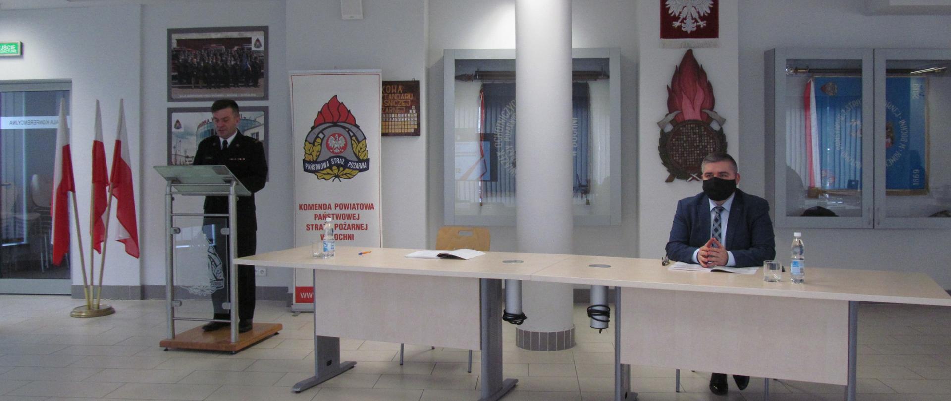 1. Duża sala konferencyjna, oświetlona światłem z lamp umieszczonych na suficie. Dwóch mężczyzn w tym jeden w mundurze wyjściowym strażackim, przemawia stojąc na mównicy ustawionej obok stołu przy którym siedzi drugi mężczyzna w garniturze z założoną czarną maseczką na twarzy, przysłuchując się przemawiającemu. W tle Godło Państwowe, trzy flagi Polski włożone w specjalny stojak, dwa sztandary zamknięte w specjalnych przeszklonych gablotach, oraz logo PSP wykonane na specjalnym rollerze