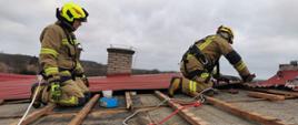 Dwóch strażaków znajduje się na dachu budynku. Są zabezpieczeni linami. Zabezpieczają uszkodzony dach.