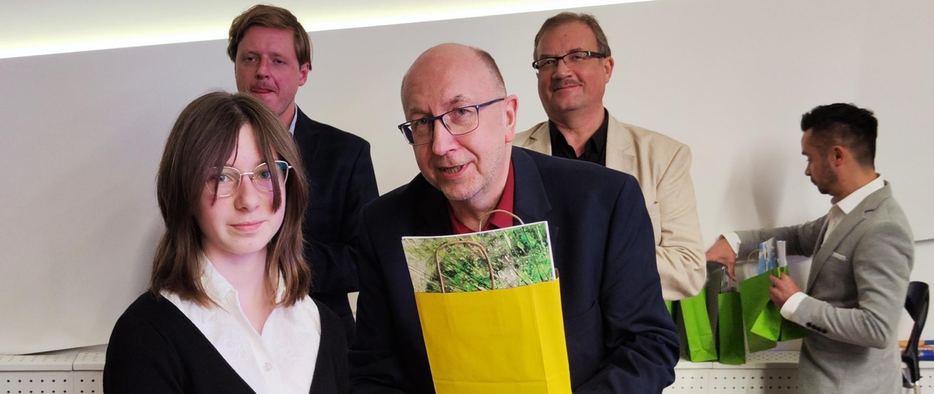 Pośrodku zdjęcia na pierwszym planie długowłosa dziewczynka w okularach ubrana na czarno podaję prawą dłoń mężczyźnie w okularach w ciemnym ubraniu, który w ręku trzyma żółtą torebkę prezentową. Z tyłu za nimi widać postacie trzech mężczyzn na jasnym tle. 