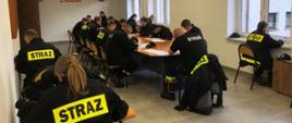 Na zdjęciu strażacy z Ochotniczych Straży Pożarnych przed egzaminem kończącym szkolenie podstawowe OSP. Strażacy ubrani w ubrania koszarowe koloru czarnego bez czapek na sali wykładowej siedzą na krzesłach przed owalnym stole i i rozwiązują testy w Komendzie Powiatowej Państwowej Straży Pożarnej w Gorlicach.