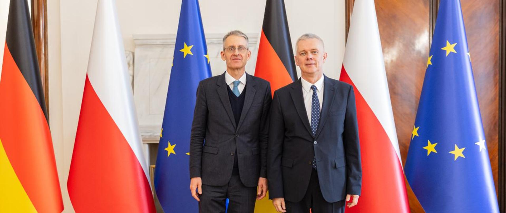 Na zdjęciu dwóch mężczyzn w eleganckich, ciemnych garniturach stoi obok siebie, przodem do obiektywu, w reprezentacyjnym wnętrzu budynku. Mężczyzna po lewej stronie ma krótkie, siwiejące włosy, okulary i krawat w odcieniu błękitu, a pod marynarką nosi ciemny sweter. Mężczyzna po prawej ma krótkie, siwe włosy, białą koszulę i granatowy krawat w drobne jasne wzory. Za nimi ustawiono naprzemiennie flagi Polski, Niemiec i Unii Europejskiej. W tle widać jasne ściany, drewniane drzwi oraz parkietową podłogę. Obaj mężczyźni stoją w formalnej pozie z rękami opuszczonymi wzdłuż ciała.
