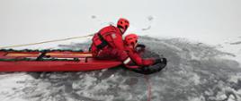 Strażak siedzący na desce lodowej podejmuje z przerębla osobę poszkodowaną, w celu bezpiecznej ewakuacji na brzeg jeziora.