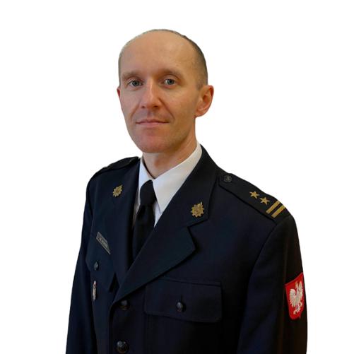 Zdjęcie portretowe mężczyzny w średnim wieku, ukazanego od pasa w górę, na czystym, białym tle. Mężczyzna jest zwrócony lekko w prawą stronę, ale patrzy bezpośrednio w obiektyw z łagodnym, profesjonalnym wyrazem twarzy. Ma jasną karnację, niebieskie oczy i jest osobą łysą.
