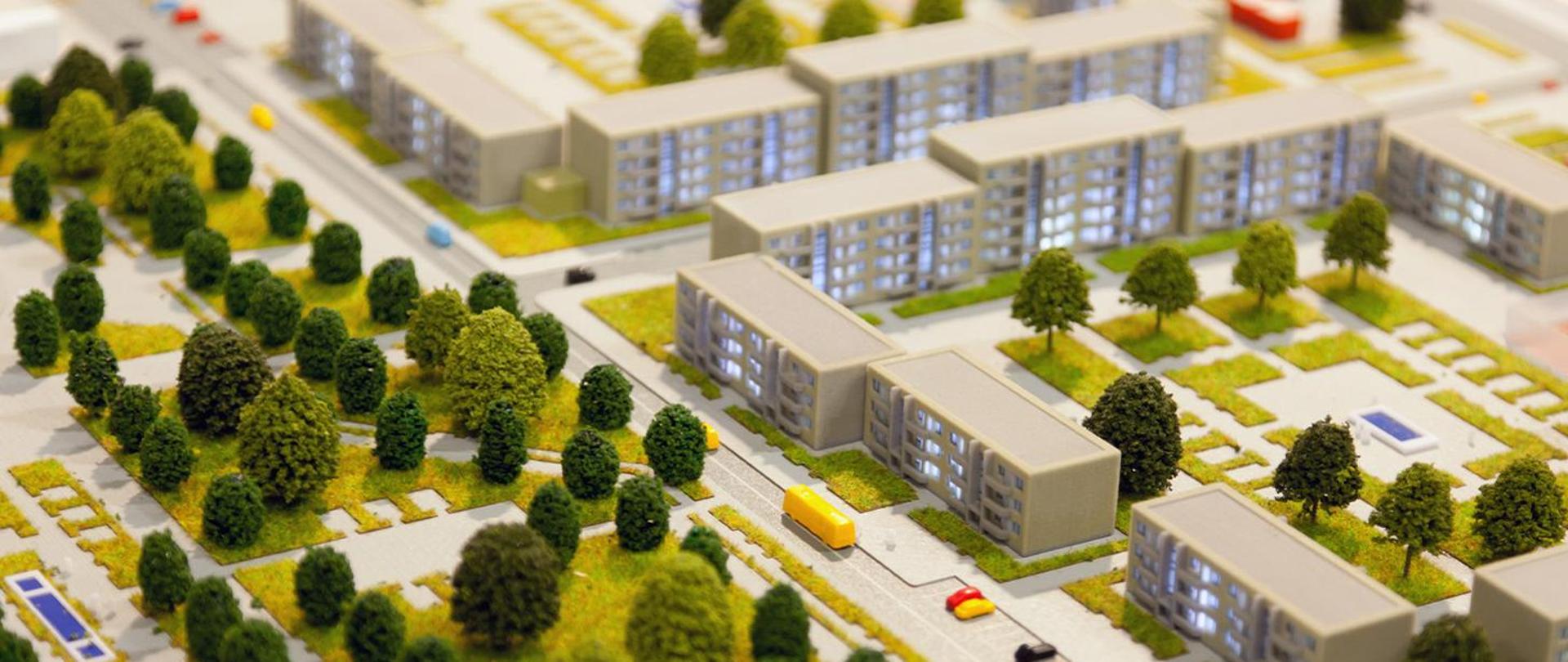 Na zdjęciu po lewej stronie znajdują się tereny zalesione. Po prawej stronie znajdują się bloki. Na dziedzińcu między blokami są drzewa. Pośrodku zdjęcia znajduje się droga, na której znajdują się pojazdy.