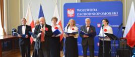 Na zdjęciu widoczna jest grupa występująca na scenie podczas oficjalnego wydarzenia. W tle znajdują się flagi Polski i Unii Europejskiej oraz niebieska ścianka z napisem Wojewoda Zachodniopomorski. Występujący są elegancko ubrani – mężczyźni w garniturach, kobiety w białych bluzkach i czarnych spódnicach, a wszyscy mają przypięte czerwone kwiaty. Trzymają w rękach teczki z nutami, a jeden z mężczyzn śpiewa lub przemawia do mikrofonu. Całość wskazuje na występ chóru lub zespołu wokalnego z okazji patriotycznej lub regionalnej uroczystości.