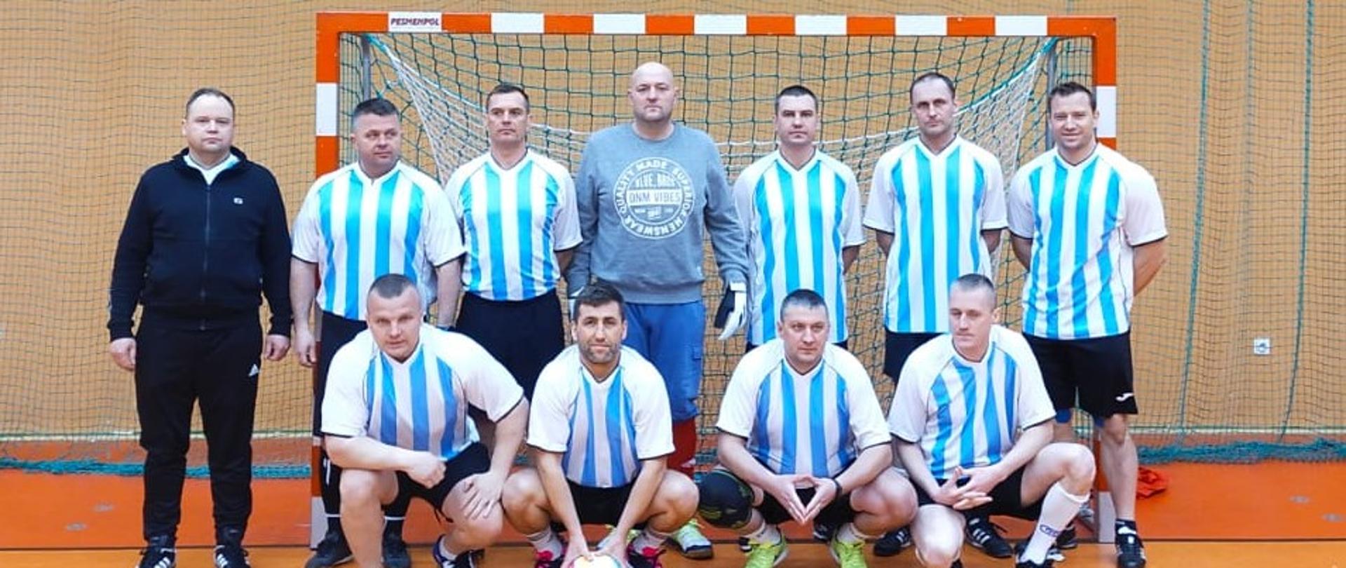 Zdjęcie zrobione na hali sportowej. Na tle bramki stoją i kuca jedenastu zawodników zawodnicy KP PSP Braniewo w biało niebieskich strojach.