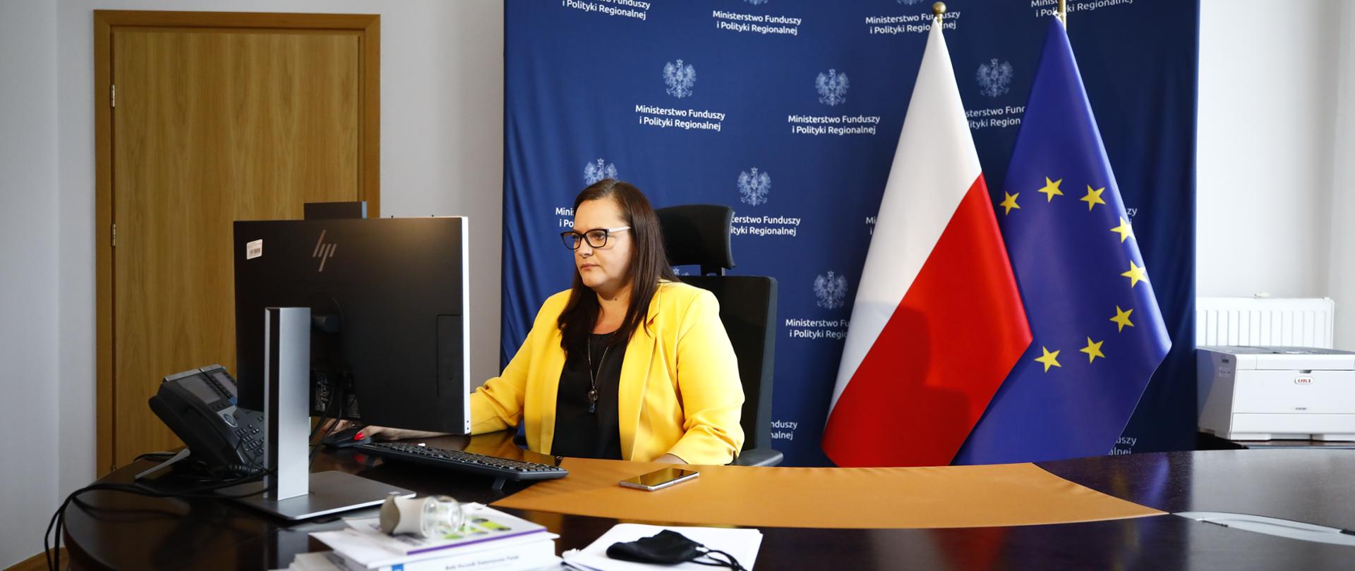 wiceminister Małgorzata Jarosińska-Jedynak uczestniczy w spotkaniu zdalnie - przed komputerem, za nią flagi Polski i Unii Europejskiej i ścianka z logotypami Ministerstwa Funduszy i Polityki Regionalnej