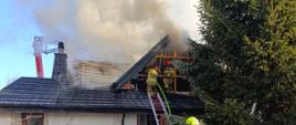 budynek mieszkalny z ciemnym dachem który u góry jest zadymiony. na dachy dwaj strażacy jeden z nich trzyma wąż na dole przed domem duża kałuża pełna wody obok przy drabinie stoi strażak 