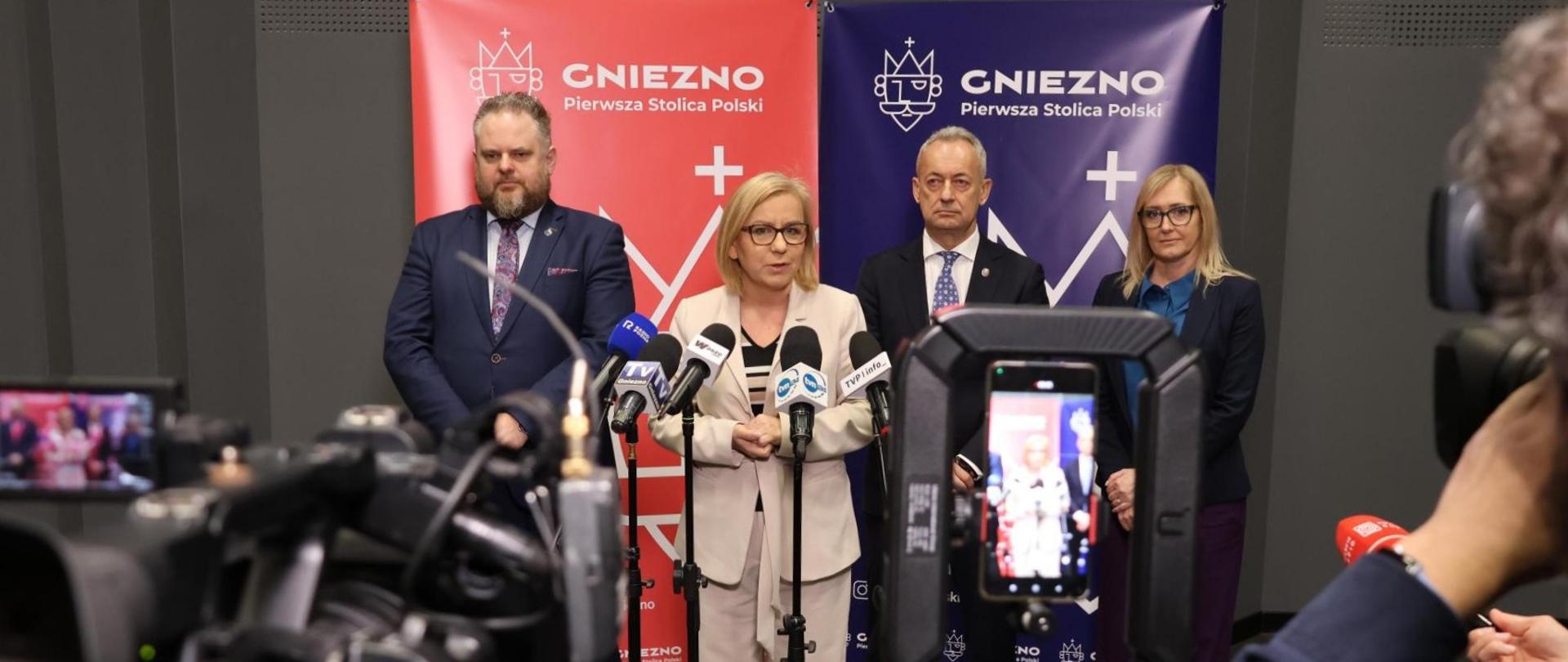 Gniezno gospodarzem ważnej europejskiej rozmowy - minister klimatu i środowiska zapowiada na konferencji spotkanie z Niemcami i Francją. Na zdjęciu ministra stoi w beżowym garniturze przed kamerami na tle 2 ścianek z czerwonym i granatowym logotypem Gniezna - Gniezno, pierwszą stolicą Polski. 