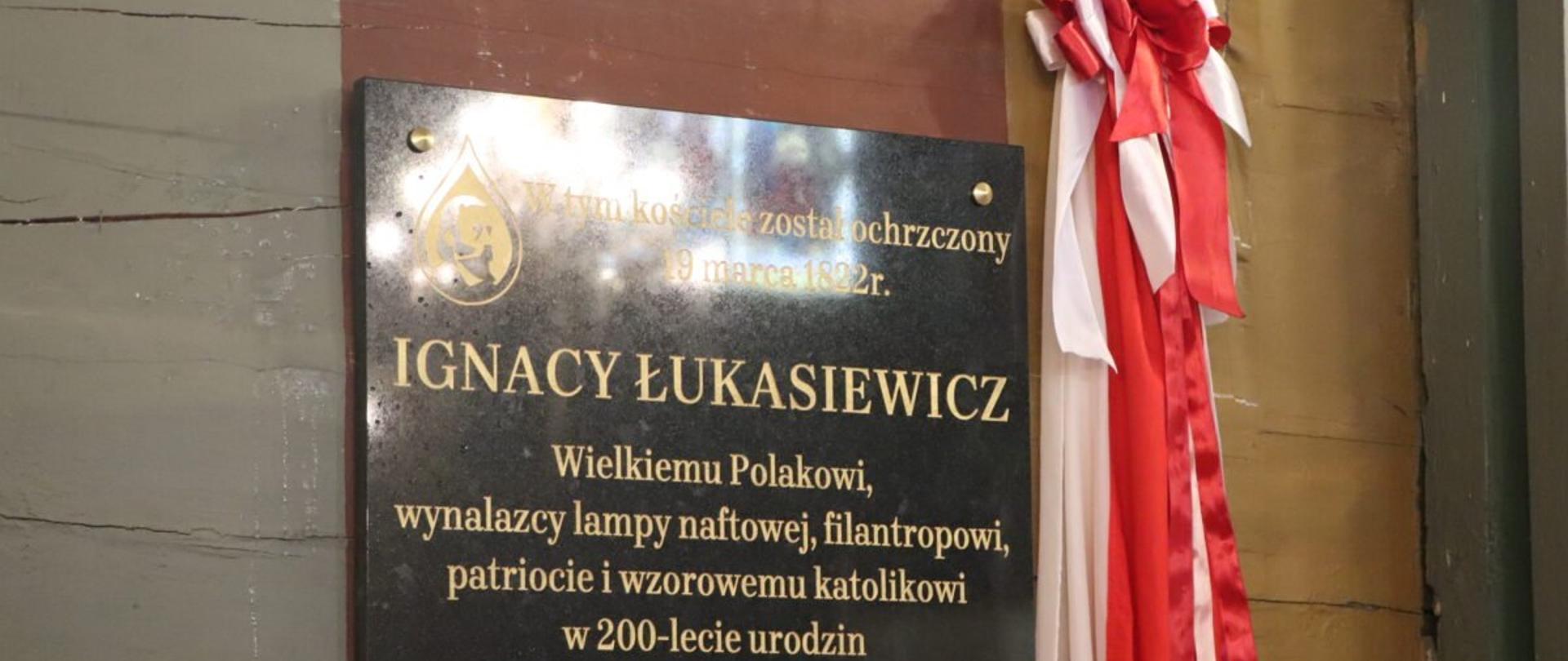 Tablica z napisem: W tym kościele został ochrzczony 19 marca 1822 Ignacy Łukasiewicz. Wielkiemu Polakowi, wynalazcy lampy naftowej, filantropowi, patriocie i wzorowemu katolikowi w 200-lecie urodzin społeczeństwo parafii Gawłuszowice, 19 marca 2022 r. Po prawej stronie tablicy widnieją wstęgi biało-czerwone