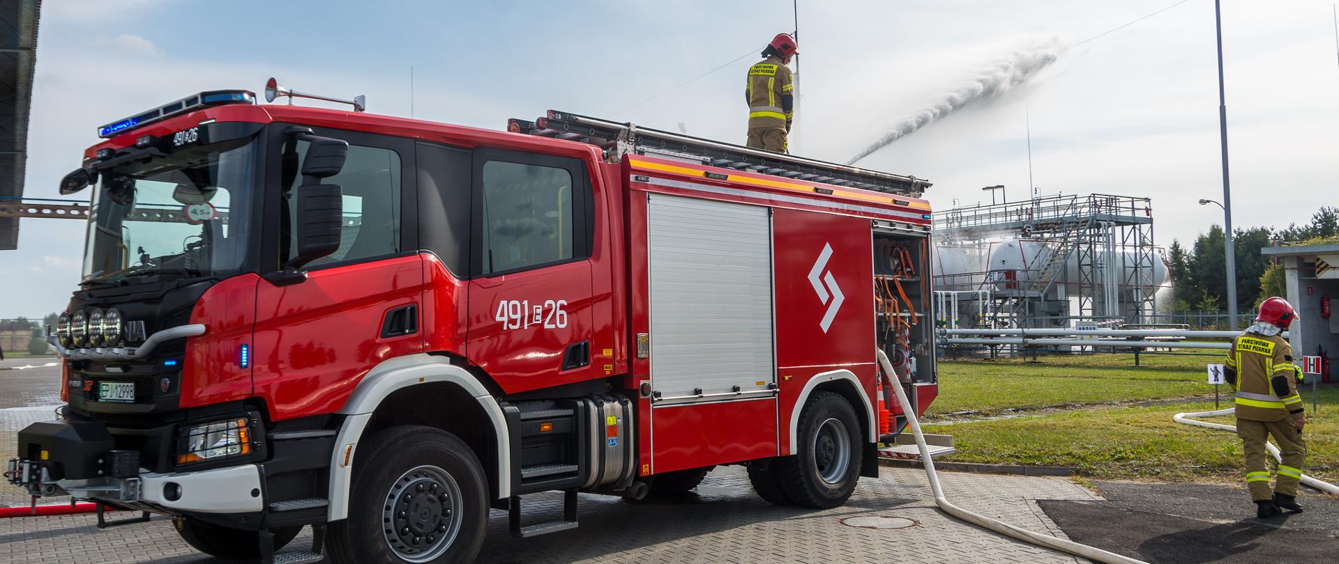 strażak podający prąd wody z działka na samochodzie gaśniczym w kierunku zbiornika z gazem, przy prawej krawędzi widać drugiego strażaka
