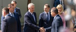 Na zdjęciu widać witających się ministra Tomasza Siemoniaka (po lewej) i jednego z węgierskich ministrów (po prawej). Po prawej i lewej stronie ministra Tomasza Siemoniaka stoją w szeregu inni ministrowie polskiego rządu. W tle widać budynek, drzewa i inne osoby.