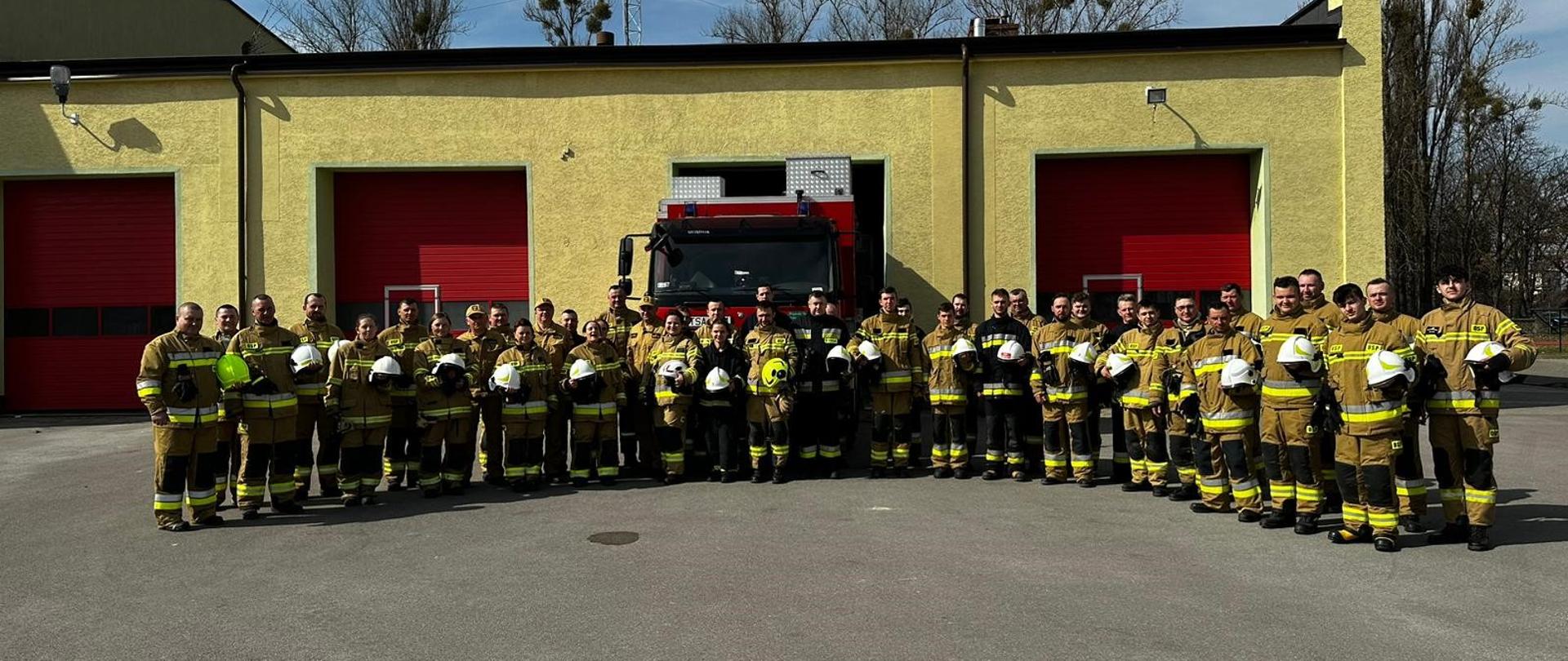 Zdjęcie przedstawia grupę 39 strażaków, którzy ukończyli kurs podstawowy dla OSP oraz w środku trzech funkcjonariuszy - oficerów PSP. Strażacy ochotnicy ubrani są w mundury bojowe, w rękach trzymają białe hełmy, natomiast mundury w kolorze piaskowy i kilku w kolorze czarnym. Strażacy ustawieni w dwuszeregu, w półkolu na tle garaży Komendy Powiatowej PSP oraz na tle czerwonego samochodu ratowniczo – gaśniczego.