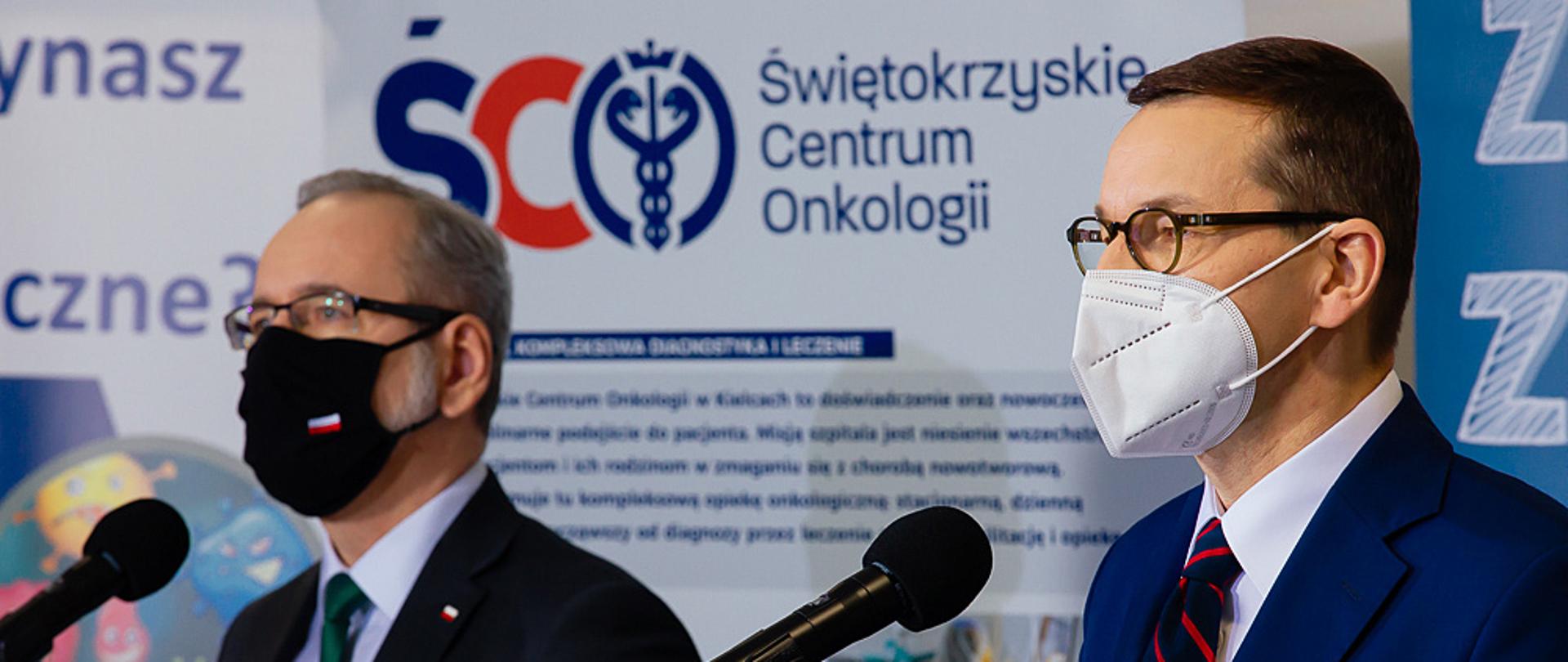 Premier Mateusz Morawiecki oraz minister Adam Niedzielski stoją przy mikrofonach podczas konferencji prasowej. W tle ścianki reklamowe Świętokrzyskiego Centrum Onkologii.