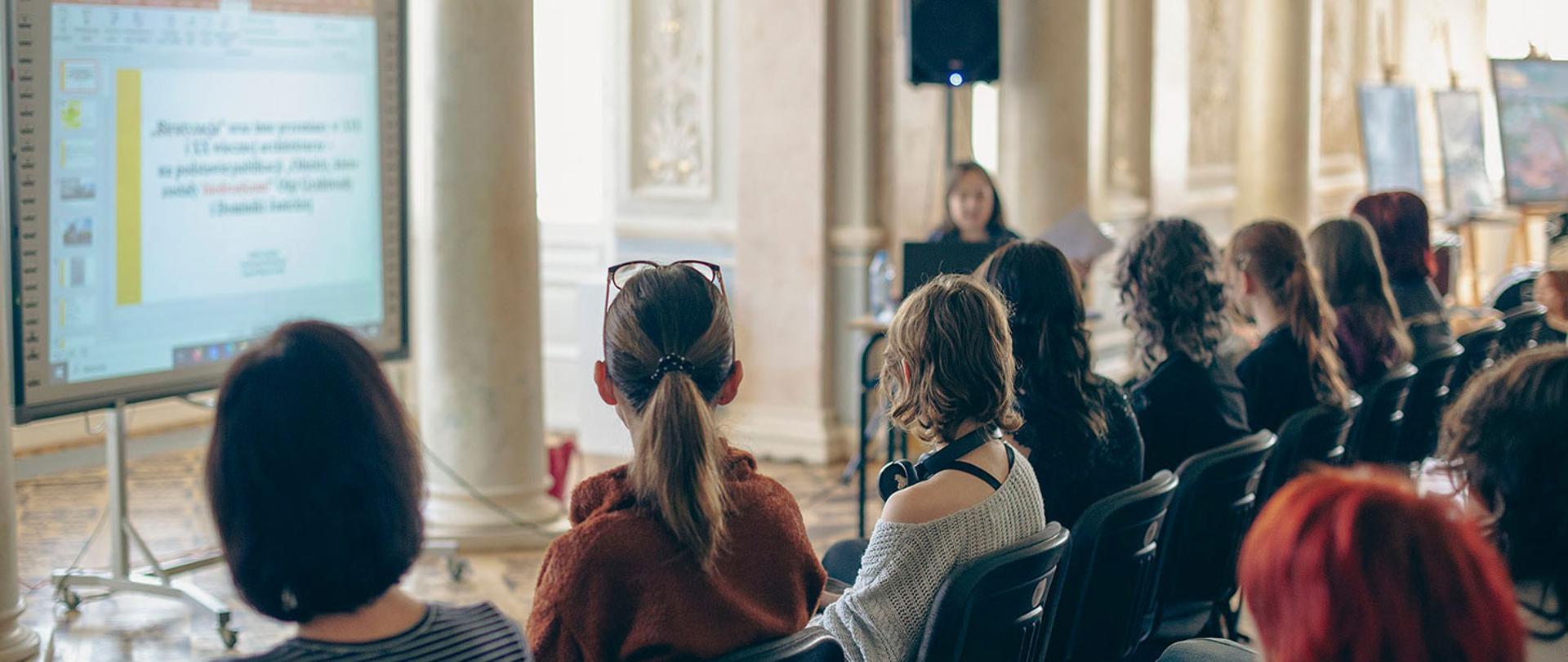 Zdjęcie przedstawia widok z tyłu na grupę osób siedzących w rzędach na czarnych krzesłach. Publiczność, składająca się głównie z młodych ludzi, patrzy w stronę dużego ekranu multimedialnego po lewej stronie. Na ekranie wyświetlany jest slajd z tekstem w języku polskim. W tle, przy stole, widać sylwetkę prelegentki prowadzącej spotkanie w dużej, widnej sali z wysokimi kolumnami.
