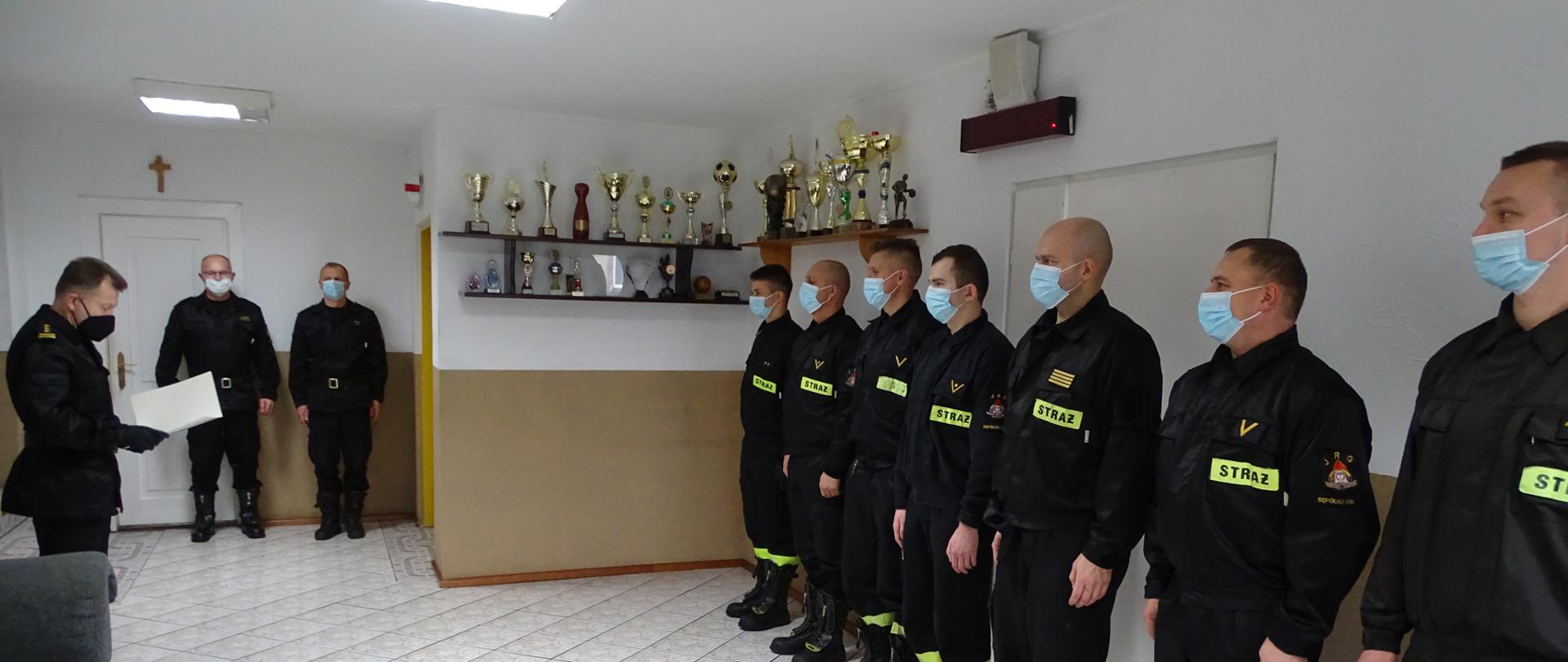 Zdjęcie wykonano w budynku Komendy Powiatowej Państwowej Straży Pożarnej w Sępólnie Krajeńskim w pomieszczeniu świetlicy. Na zdjęciu po lewej stronie ubrany w mundur służbowy Komendant Powiatowy starszy brygadier Maciej Suchomski otrzymuje odczytuje rozkaz nadania awansu. Po prawej stronie strażacy ze zmiany służbowej stoją w szeregu ubrani w mundury koszarowe. W głębi po lewej stronie Zastępca Komendanta Powiatowego młodszy brygadier Dariusz Chylewski oraz dowódca Jednostki Ratowniczo Gaśniczej starszy kapitan Robert Liss ubrani w mundury służbowe.
