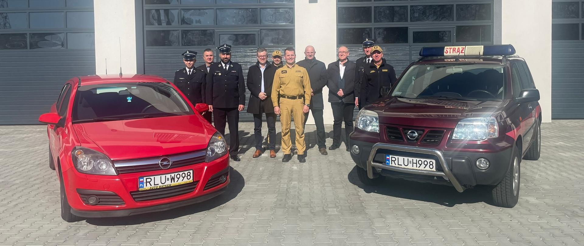 Zdjęcie przedstawia dwa samochody operacyjne na tle garaży. W centralnej części zdjęcia widoczni są ludzie ubrani w mundury OSP, garnitury oraz piaskowe mundury PSP.