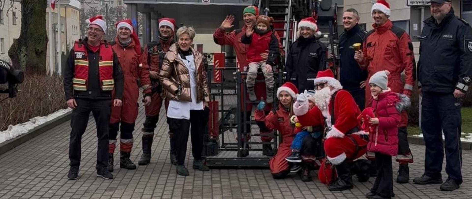 Grupa organizatorów akcji - strażacy i policjanci stojący na placu z kostki brukowej przed szpitalem