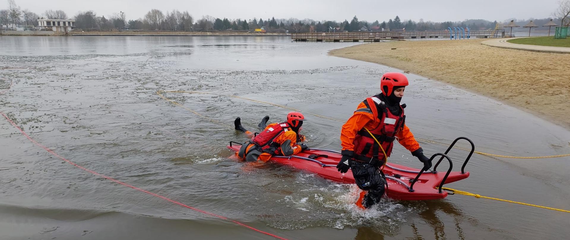 Strażak wychodzi z wody ciągnąc obok siebie sanie lodowe, na saniach ciągniętych jest dwóch strażaków, którzy częściowo zanurzeni są w wodzie.
