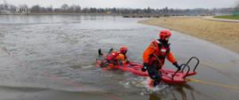Strażak wychodzi z wody ciągnąc obok siebie sanie lodowe, na saniach ciągniętych jest dwóch strażaków, którzy częściowo zanurzeni są w wodzie.