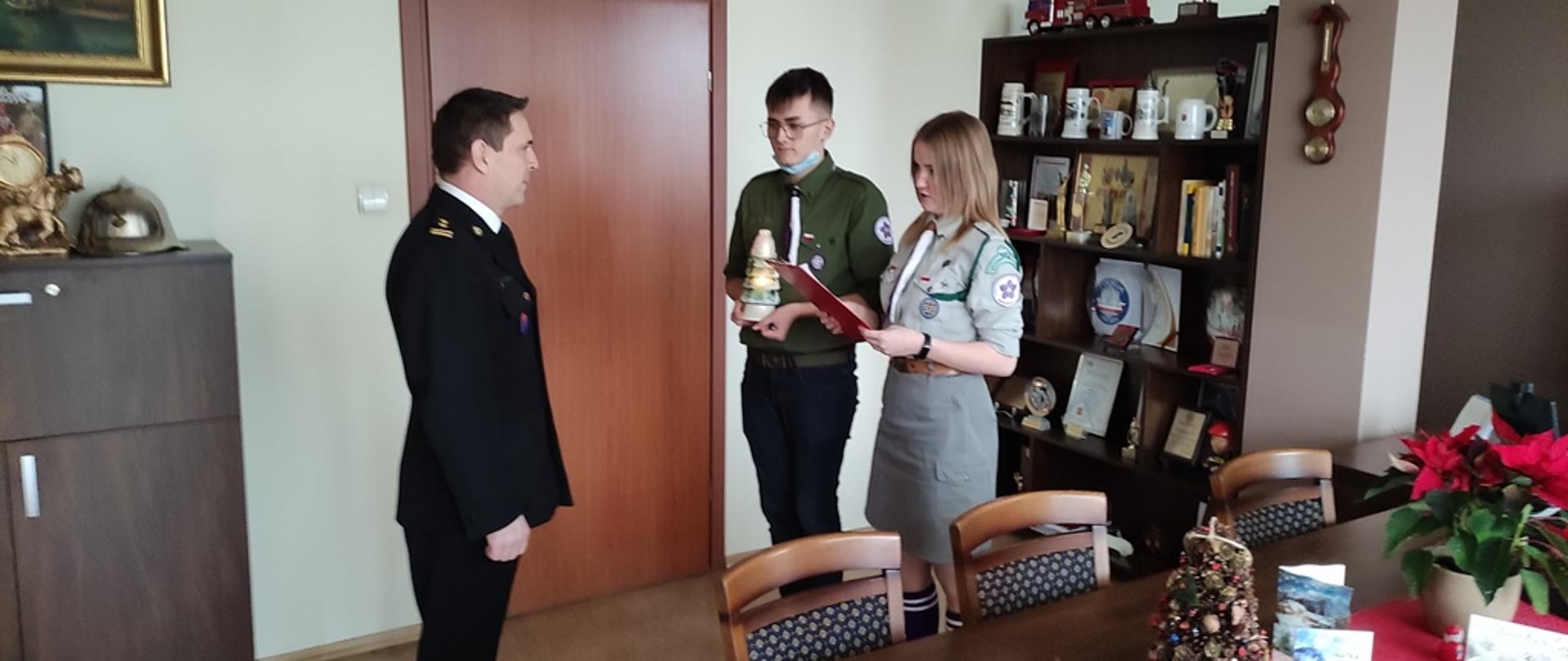 Gabinet komendanta powiatowego, z lewej strony w ubraniu wyjściowym zastępca komendanta powiatowego PSP, z prawej strony harcerka i harcerz, w trakcie odczytywania tegorocznego przesłania i hasła akcji, za harcerzami gablota z pamiątkowymi statuetkami okolicznościowymi