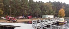Widok na pomost na jeziorze a w tle pojazdy strażackie oraz pogotowia ratunkowego stoją nad brzegiem jeziora.