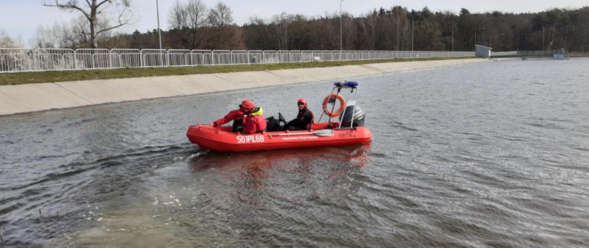 Trzech strażaków w ubraniach ochronych, kaskach i kamizelkach ratunkowych płynie po jeziorze w czerwonej łodzi motorowej. w tle widoczna tama i las