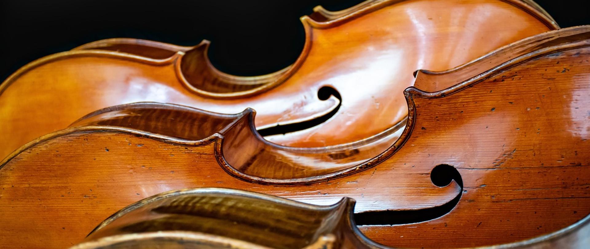 Trzy ułożone za sobą wiolonczele. Widoczne tylko górne części pudeł rezonansowych. Czarne tło