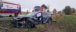 W przydrożnym rowie stoi samochód z uszkodzonym przodem. Na jezdni stoi samochód osobowy, karetka pogotowia ratunkowego oraz samochody strażackie. Na jezdni porozrzucane plastiki. 