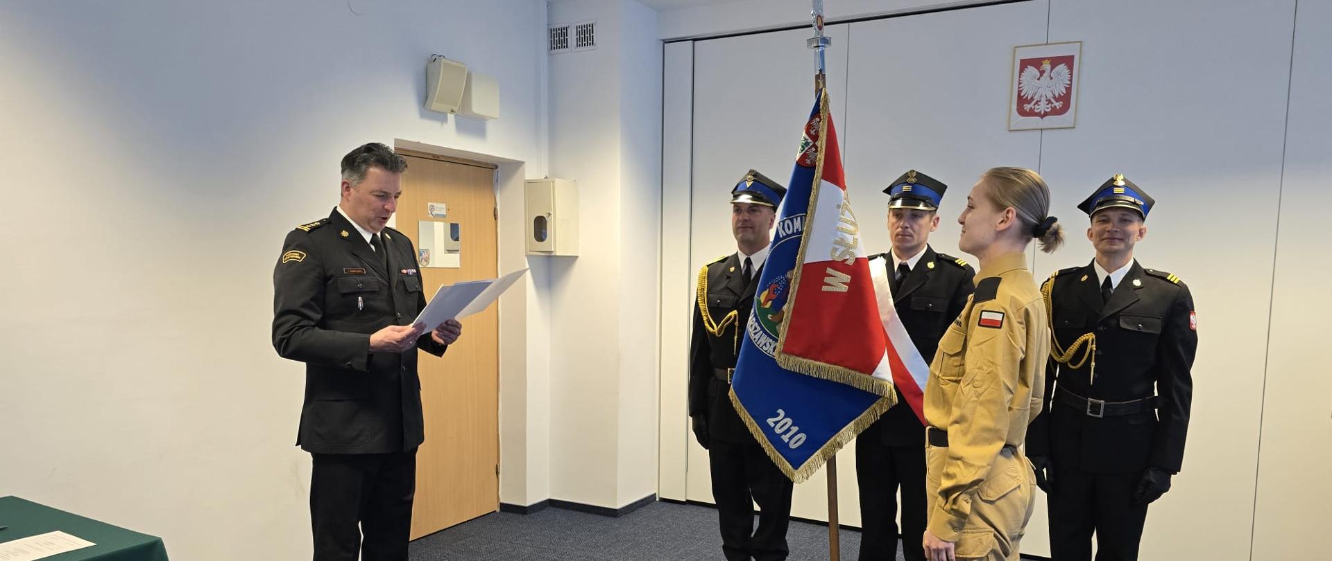 Komendant w stroju wyjściowym czyta akt powołania nowego strażaka który stoi na przeciwko a w tle poczet sztandarowy. na scianie wisi Godło Polski