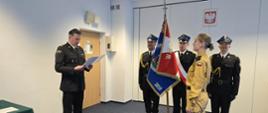 Komendant w stroju wyjściowym czyta akt powołania nowego strażaka który stoi na przeciwko a w tle poczet sztandarowy. na scianie wisi Godło Polski