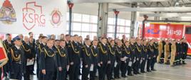 Na zdjęciu widoczna jest uroczystość w jednostce straży pożarnej.
Na pierwszym planie stoi duża grupa strażaków w galowych mundurach – są to funkcjonariusze Państwowej Straży Pożarnej, co widać po ich czarnych mundurach z charakterystycznymi złotymi sznurami.
Po lewej stronie widać fragment mównicy i mikrofonu, co sugeruje, że uroczystość zawiera oficjalne przemówienia.
W tle znajduje się baner z logotypem KSRG (Krajowy System Ratowniczo-Gaśniczy) oraz herbami Państwowej Straży Pożarnej i innych służb pożarniczych.
W tle znajduje się także nowoczesny wóz strażacki z oznaczeniem "341[N]50".
Całość ma miejsce w hali garażowej strażnicy, która została specjalnie przygotowana na tę uroczystość – widać czystość, porządek i brak pojazdów na stanowiskach poza jednym.
Zdjęcie przedstawia więc formalne wydarzenie związane z obchodami Dnia Strażaka, wręczeniem odznaczeń i awansów.