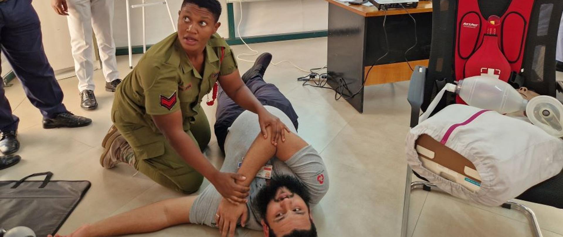 Mężczyzna leży na ziemi, kobieta w mundurze pokazuje na nim zasady pierwszej pomocy