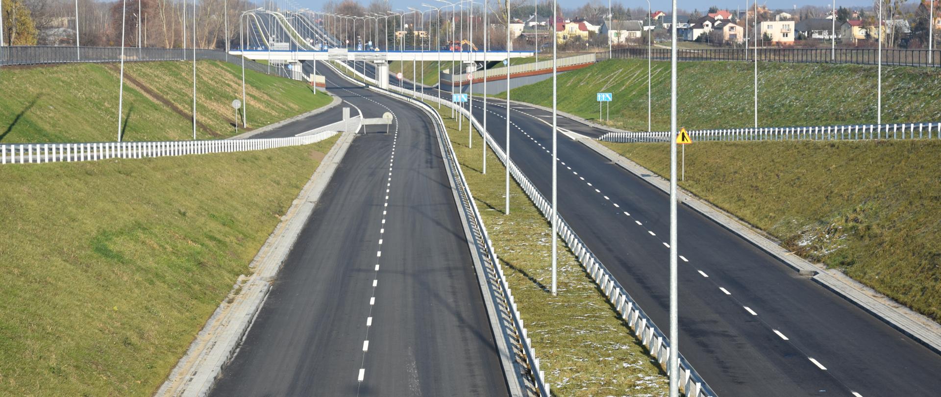 Na zdjęciu widać fragment przebudowanej drogi. Pomiędzy jezdniami znajduje się pas zieleni z latarniami. Po bokach jezdni również znajduje się pas zieleni.