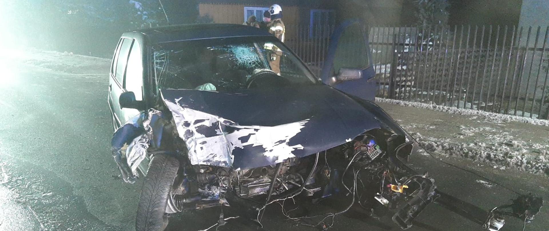 Na drodze asfaltowej stoi samochód osobowy, koloru granatowego z prawie całkowicie skasowanym przodem w wyniku wypadku. Za samochodem widać dom jednorodzinny, ogrodzony oraz stojących przy im ludzi: strażaka OSP oraz jednego mężczyznę. 