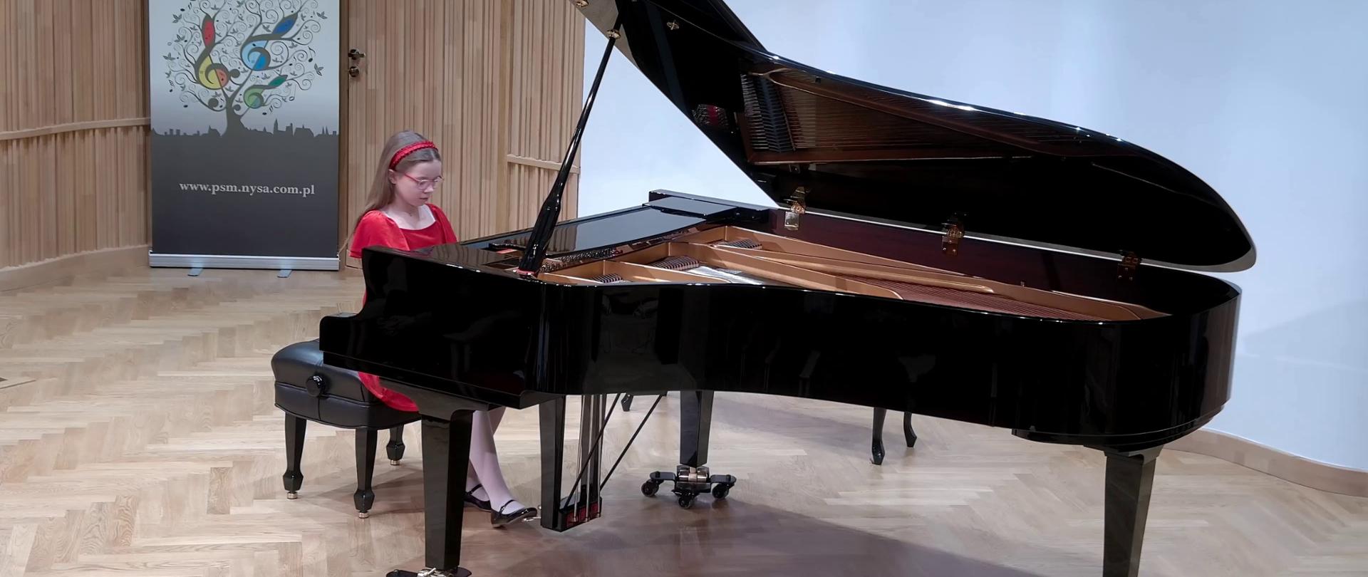 Zdjęcie. Na estradzie Sali Koncertowej stoi czarny duży fortepian, na którym gra uczennica ubrana w czerwona sukieneczkę. W tle stoi baner PSM Nysa.