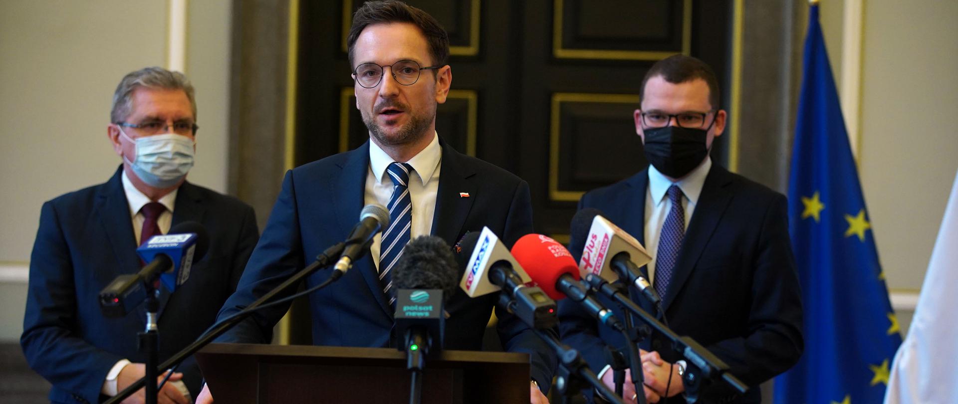 Trzech mężczyzn przed mikrofonami. W środku stoi wiceminister Waldemar Buda.