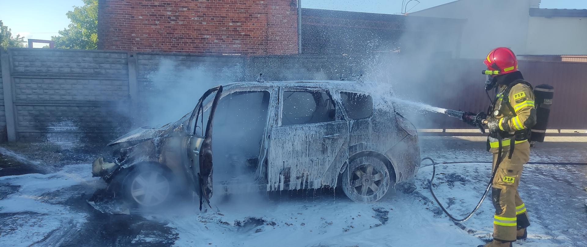 Na zdjęciu spalony samochód. Widać środek gaśniczy oraz ratownika w ubraniu specjalnym Zdjęcie w ciągu dnia. Ratownik posiada hełm. Z tyłu zabudowania.