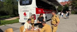 Funkcjonariusze Komendy wraz z uczestnikami akcji pobrania krwi na tle autobusu wypełniający dokumentację. Osoby siedzą przy stolikach na placu wewnętrznym komendy.