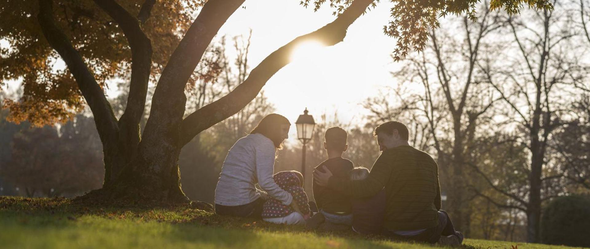 Rodzice siedzący z dziećmi na trawie w otoczeniu drzew 