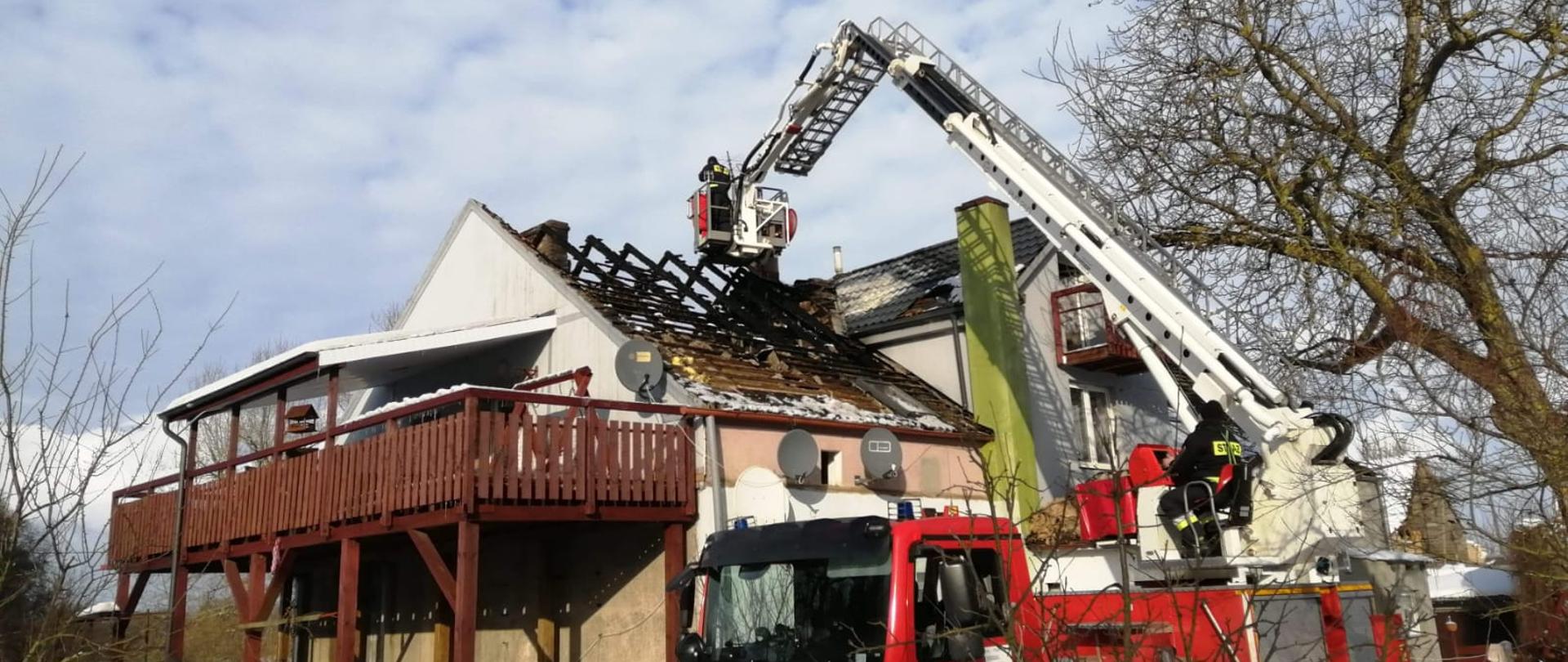 Zdjęcie przedstawia budynek wielorodzinny ze spalonym dachem w porze zimowej. Przed budynkiem znajduje się wóz strażacki z podnośnikiem koszowym z którego strażacy przykrywają dach.