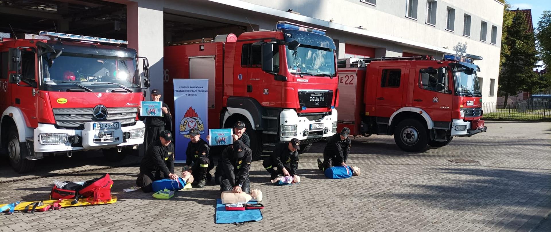 Zdjęcie przedstawia funkcjonariuszy Komendy Powiatowej Państwowej Straży Pożarnej w Lęborku podczas prowadzenia resuscytacji krążeniowo- oddechowej na fantomach ćwiczebnych. W tle ustawione pojazdy ratowniczo-gaśnicze.