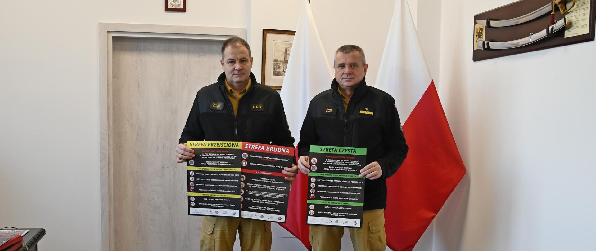Funkcjonariusze Państwowej Straży Pożarnej stoją trzymając plakaty za nimi stoją flagi Polski oraz na ścianie wisi szabla.