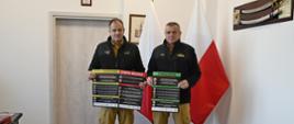 Funkcjonariusze Państwowej Straży Pożarnej stoją trzymając plakaty za nimi stoją flagi Polski oraz na ścianie wisi szabla.